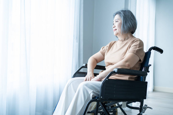 Ασιάτισσα μεγαλύτερη ώριμη γυναίκα κάθεται μόνη σε αναπηρικό καροτσάκι στο σπίτι. Μοναχικές ηλικιωμένες γυναίκες Η γιαγιά ασθενής είναι ευτυχισμένη και απολαμβάνει την ανατολή κατά τη διάρκεια του πρωινού στο γηροκομείο. Νοσοκομειακή περίθαλψη και ιατρική. - Φωτογραφία, εικόνα
