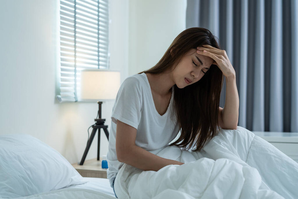 Asian depressed woman feel headache sitting alone on bed in bedroom. Attractive unhappy young sick girl feeling sad lonely and upset with life problem then crying with tears in dark night room at home - Photo, image