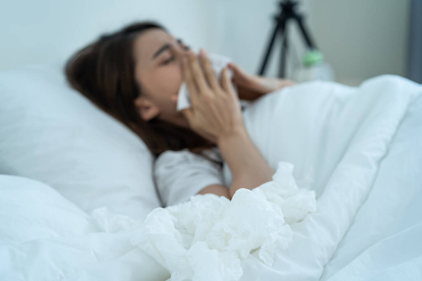 Asian sick girl in pajamas wake up from sleep at night sneezing on bed. Attractive young woman feeling bad and suffer from allergy, put tissue cover her nose while sneezes during bedtime in house. - Photo, image
