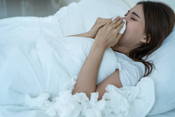 Asian sick girl in pajamas wake up from sleep at night sneezing on bed. Attractive young woman feeling bad and suffer from allergy, put tissue cover her nose while sneezes during bedtime in house. - Photo, image