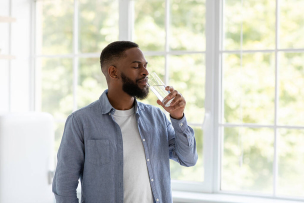 Sakallı yakışıklı siyahi adam pencerenin arkasında kapalı gözlerle temiz su içiyor. - Fotoğraf, Görsel