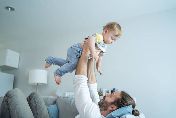 Caucasian loving dad play with baby boy child on sofa in living room. Happy family, attractive handsome caring young father lifting toddler son fly up enjoy doing airplane game in morning in house. - Zdjęcie, obraz