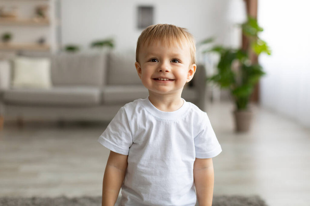Portrét roztomilého batolete chlapce v bílém tričku a s úsměvem, pózování na kameru v interiéru obývacího pokoje - Fotografie, Obrázek