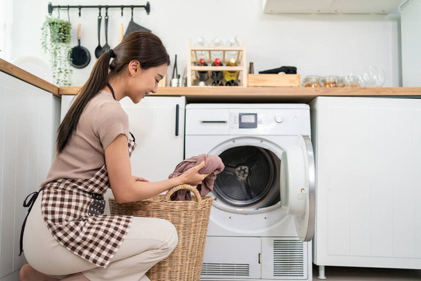 Aziatische mooie vrouw zet vuile kleren aan wasmachine in huis. Aantrekkelijk meisje dragen schort zitten op de vloer, voel me blij om de was te laden in wasmachine apparaat thuis. Huishoudconcept. - Foto, afbeelding