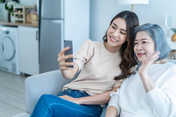 Aasialainen onnellinen perhe, vanhempi äiti käyttää matkapuhelinta tyttärensä kanssa kotona. Houkutteleva kaunis tyttö istua vanhusten kanssa vanhempi äiti sohvalla ja video puhelun verkossa kommunikoida chat olohuoneessa talossa. - Valokuva, kuva