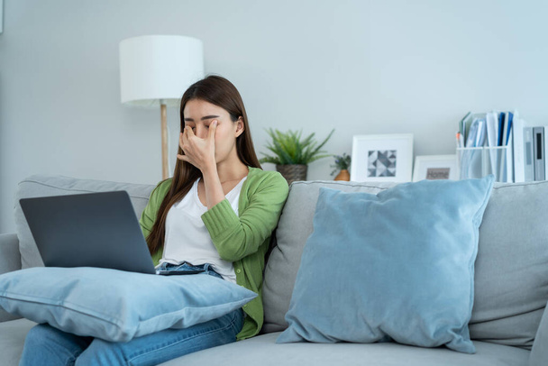 Exhausted Asian business woman hurt eye while using laptop computer. Young girl office worker sit on sofa feel visual fatigue and eye strain tired from overwork and massage dry irritable eye at home. - 写真・画像