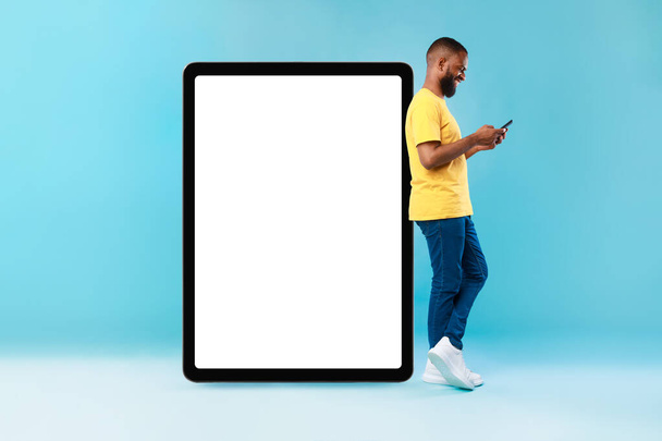 Ελκυστικός μαύρος άνδρας στέκεται κοντά σε τεράστια ψηφιακή ταμπλέτα με άδεια οθόνη, χρησιμοποιώντας smarthone, διαφημίζοντας νέα εφαρμογή, mockup - Φωτογραφία, εικόνα