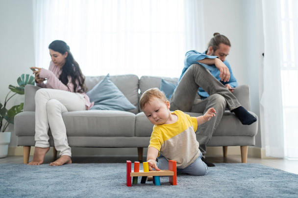 Caucasian phone addict parents don't pay attention with baby toddler. Family problem, Father and Mother sit on sofa and using cellphone ignore young infant boy son play alone on floor in living room. - 写真・画像