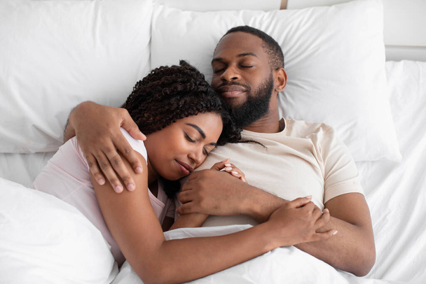 Ευτυχισμένος νεαρός μαύρος σύζυγος αγκαλιάζει τη γυναίκα του και ξαπλώνει στο κρεβάτι πάνω σε μαλακό μαξιλάρι κάτω από λευκή κουβέρτα στην κρεβατοκάμαρα. - Φωτογραφία, εικόνα