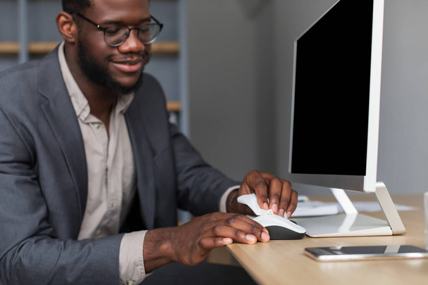 Millennial black businessman sitting ar desk with pc monitor, wiping computer mouse with sanitizer at office, mockup - Valokuva, kuva