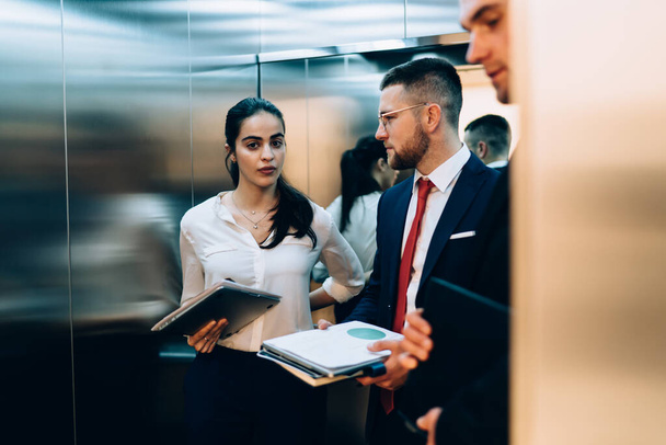 Serious group of young business people standing next to each other in elevator and carrying files and documents in hands while wearing formal outfit - Fotografie, Obrázek