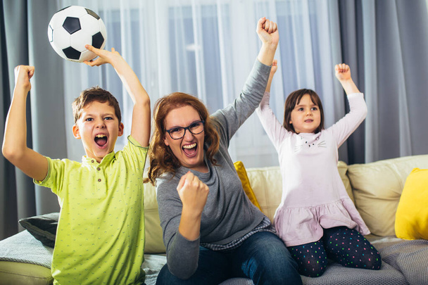Mother and her kids raising arms and screaming while watching football match and celebrating goal at hom - Foto, Imagem