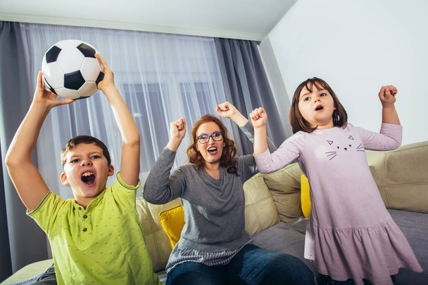Madre y sus hijos levantan los brazos y gritan mientras ven el partido de fútbol y celebran el gol en hom - Foto, Imagen