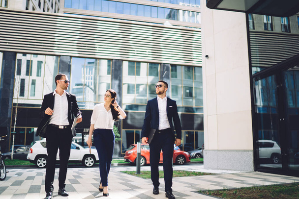 Full length of busy young coworkers in formal elegant clothes strolling along contemporary business centre and discussing about team work - Foto, imagen