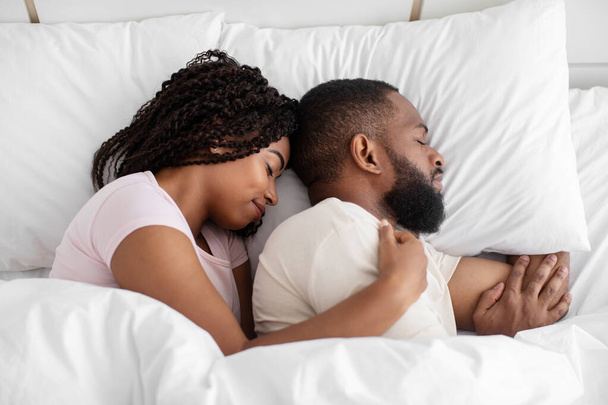 Milenyum siyahi kadın ve erkek, yatak odasında beyaz battaniyenin altında, yumuşak yastığın üzerinde uzanıp, uyuyup, sarılıyorlar. - Fotoğraf, Görsel