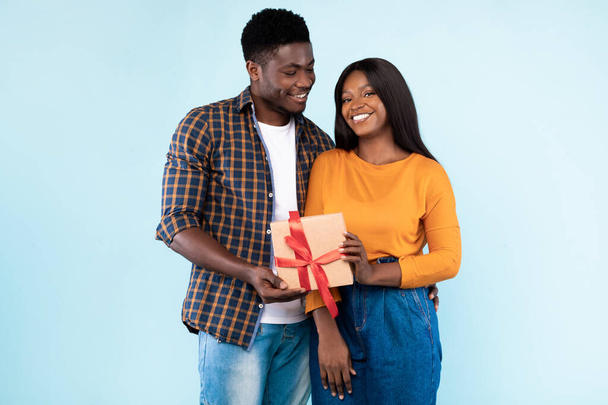 Happy black couple holding gift box at blue studio - Zdjęcie, obraz