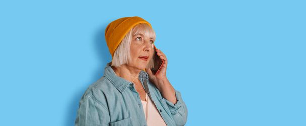 Οριζόντια λήψη χαρούμενα σύγχρονη ηλικιωμένη γυναίκα χρησιμοποιεί το τηλέφωνο μιλάμε σε ένα κινητό τηλέφωνο έχει χαρούμενη διάθεση πάνω από το μπλε φόντο χρώμα - Φωτογραφία, εικόνα