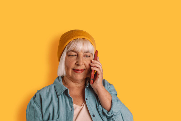 Portrait of 50s blonde woman holds smartphone near ear posing uses internet for communication against yellow wall background. Modern technology and communication - Fotografie, Obrázek