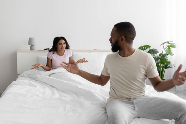 Unhappy angry excited millennial african american wife and husband swears and yells on bed in white bedroom - Fotó, kép