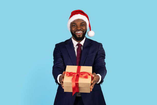 Emocionado hombre de negocios negro con sombrero de Santa dar caja de regalo en la cámara - Foto, imagen