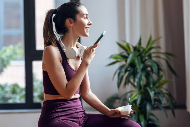 Shot of sporty young woman eating a yogurt while using smartphone sitting on fitness ball at home. - Foto, Imagem