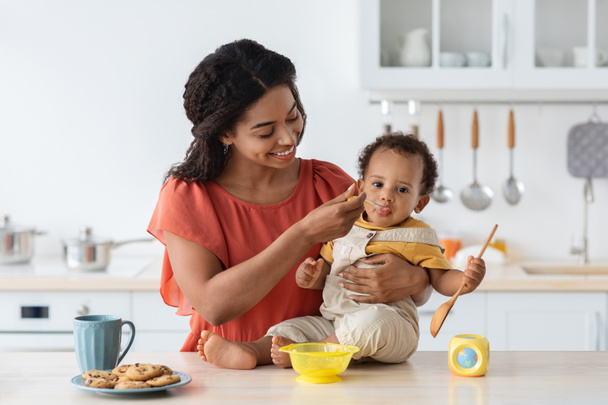 Vauvan vieroitus. Huolehtiva musta äiti ruokinta lapsi poika lusikka keittiössä - Valokuva, kuva