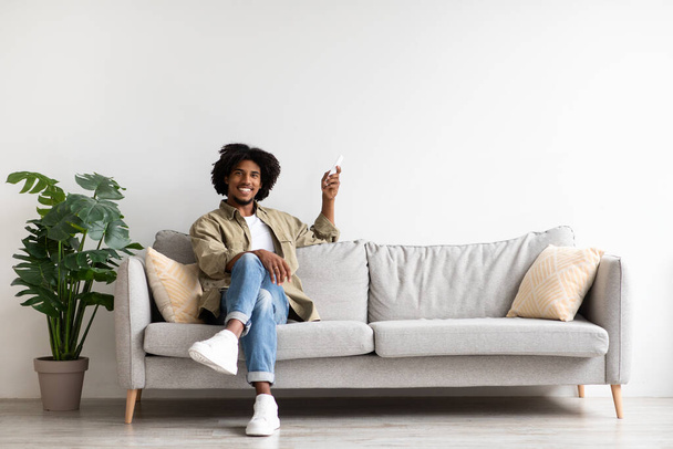 Home Comfort. Jeune homme noir assis sur le canapé et tenant télécommande - Photo, image