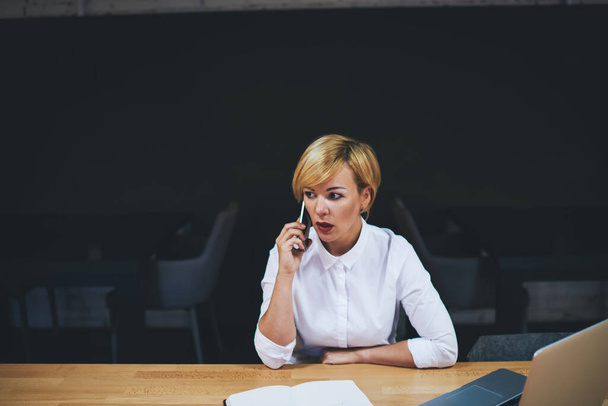 Young concentrated blond female manager in formal clothes having conversation on mobile phone sitting at table with laptop in office - Foto, Imagem