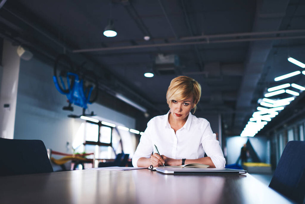 Confident female employee in white shirt looking at camera while sitting at table in modern workplace and writing notes during workday - Foto, Imagen