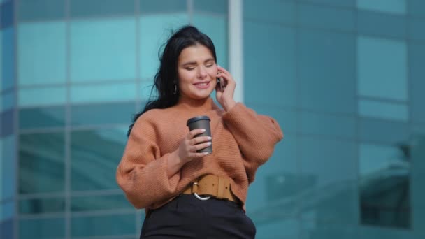 Young hispanic woman standing outdoors friendly talking on mobile phone cute curvy girl drinking hot coffee businesswoman resting at lunchtime pleasant conversation telephone orders using smartphone - Footage, Video
