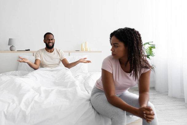 Δυστυχισμένος θυμωμένος νεαρός μαύρος σύζυγος φρικάρει και φωνάζει στη θλιμμένη σύζυγο, κάθεται στο κρεβάτι - Φωτογραφία, εικόνα
