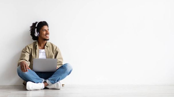 Cheerful Black Guy In Wireless Headphones Relaxing With Laptop On Floor - Foto, Imagen