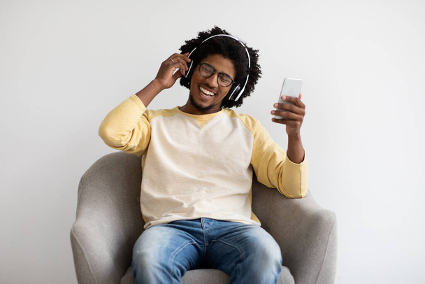 Νεαρός μαύρος τύπος με Smartphone και ασύρματα ακουστικά χαλαρώνοντας στην πολυθρόνα - Φωτογραφία, εικόνα