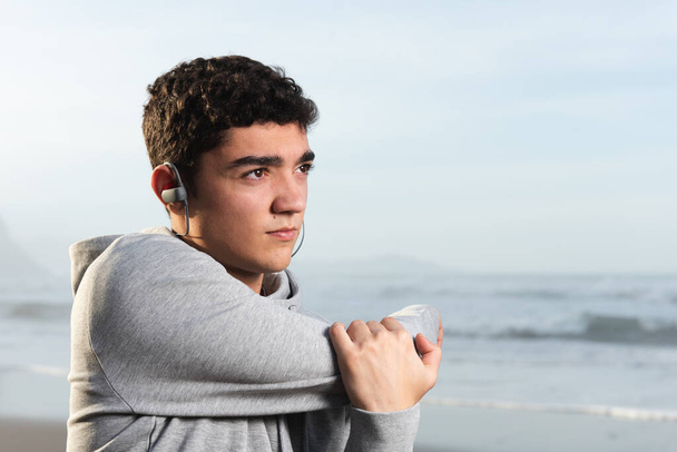 Retrato de adolescente hispano estirándose y calentándose mientras escucha un podcast en sus auriculares. - Foto, Imagen