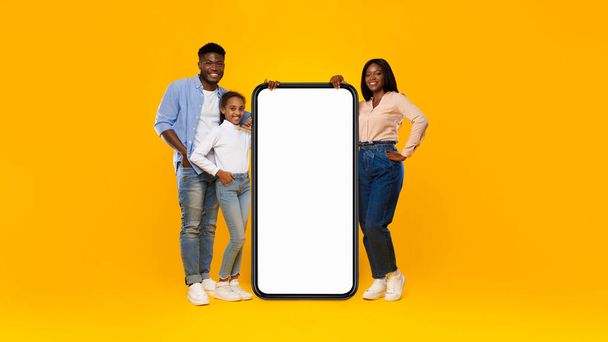 Black family presenting white empty smartphone leaning on big screen - Фото, изображение