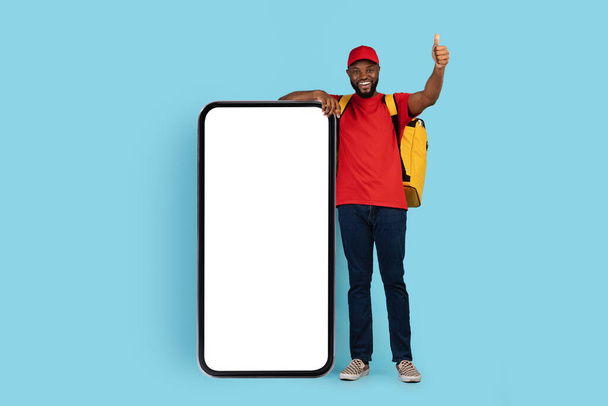 Happy African American Delivery Guy With Thermal Backpack Standing Near Blank Smartphone - Φωτογραφία, εικόνα