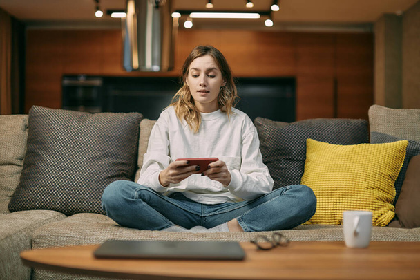 Female gamer playing mobile online game on smart phone with friends online sitting on sofa at home living room - Foto, immagini