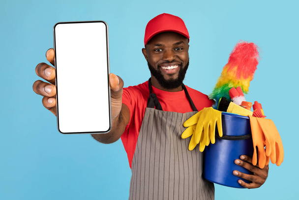 Black male cleaner holding basket with cleaning tools and showing blank smartphone - Foto, Bild