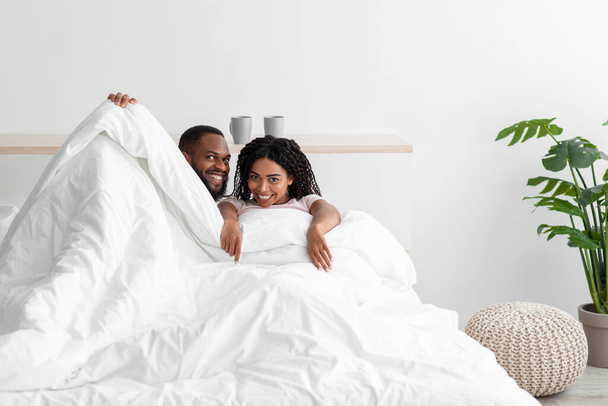 Yatakta battaniyeye sarılı gülümseyen genç siyahi kadın ve erkek haftasonunun keyfini çıkarın. - Fotoğraf, Görsel
