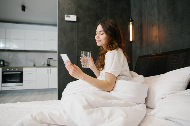Relaxed young woman drinking water and using cell phone in bed - stock photo - Photo, image