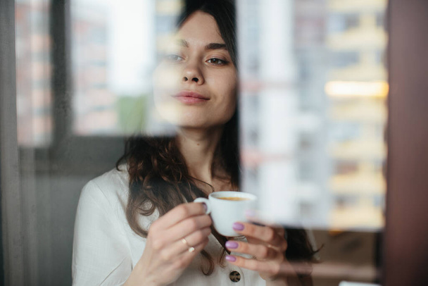 Beautiful caucasian woman drinking hot coffee or tea and looking through window. Indoor background. - Foto, Imagem