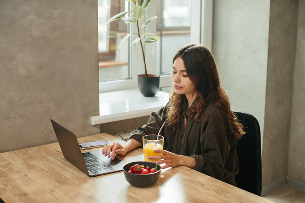 Le portrait d'une belle jeune femme travaillant avec un ordinateur portable pendant le petit déjeuner avec des fraises et boire du jus d'orange. Manger à la maison. - Photo, image