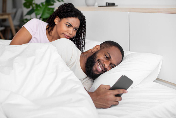 Αναστατωμένη μαύρη γυναίκα χιλιετηρίδας κοιτάζει το smartphone του συζύγου της, κουβεντιάζοντας στο τηλέφωνο στα κοινωνικά δίκτυα και να παίξει - Φωτογραφία, εικόνα