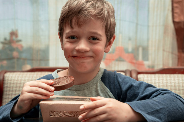 portrait of a funny cute Caucasian boy who eats chocolate sitting at a table in a home kitchen - Foto, Imagen