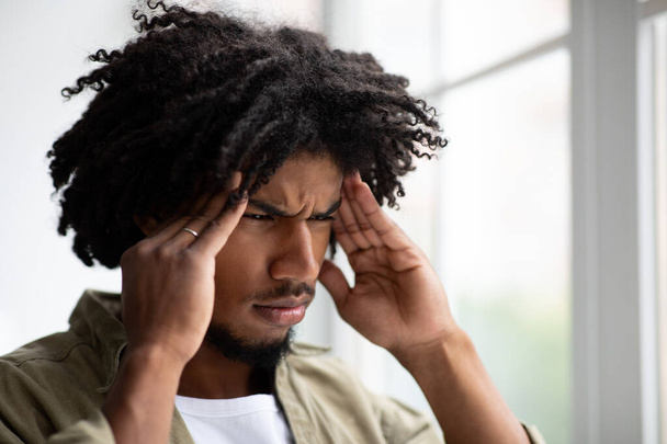 Αναστατωμένος νεαρός μαύρος που υποφέρει από πονοκέφαλο ενώ στέκεται κοντά στο μεγάλο παράθυρο - Φωτογραφία, εικόνα