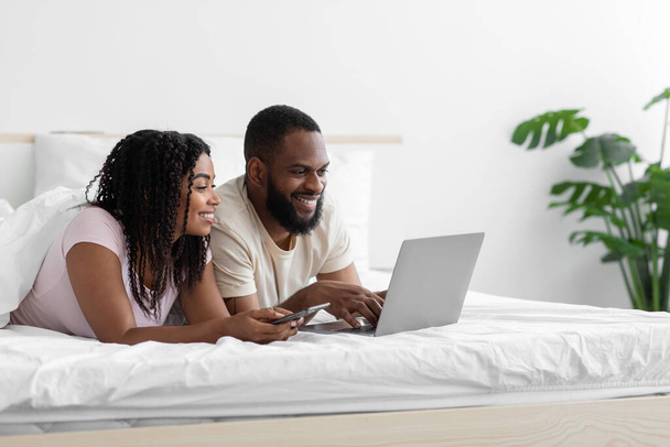 Happy young black husband and wife lie on bed and watch video on computer in free time - Foto, imagen