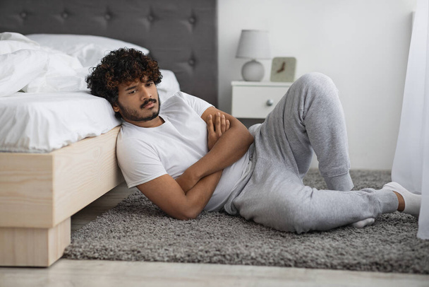 Pensive indian guy in pajamas sitting on floor by bed - 写真・画像