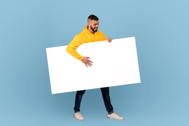 Excited arab man walking with white blank placard under arm over blue studio background, mockup for text or design - Photo, Image