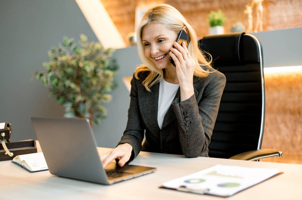 Mature happy joyful confident caucasian blonde business lady, broker or manager, sitting at table in a modern office, working with laptop, talking to client or employees on smartphone, smiling - Φωτογραφία, εικόνα
