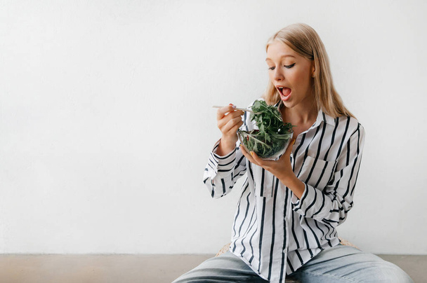Healthy lifestyle concept. Young blonde woman sitting on the floor, eating salad and smiling - Photo, image
